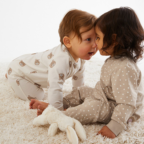 Två toddlers sitter på en fluffig matta klädda i mönstrad babypyjamas